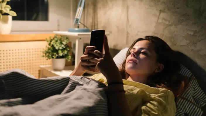 Frau liegt im Bett und schaut auf ihr Smarthone. 