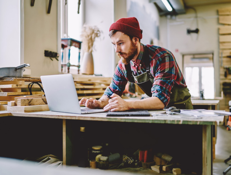 Handwerker sitzt in seiner Werkstatt am Laptop. 