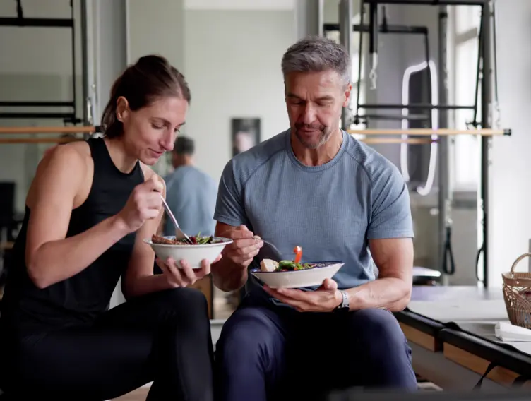 Eine Frau und ein Mann sitzen zusammen in Sportkleidung und essen etwas gesundes.