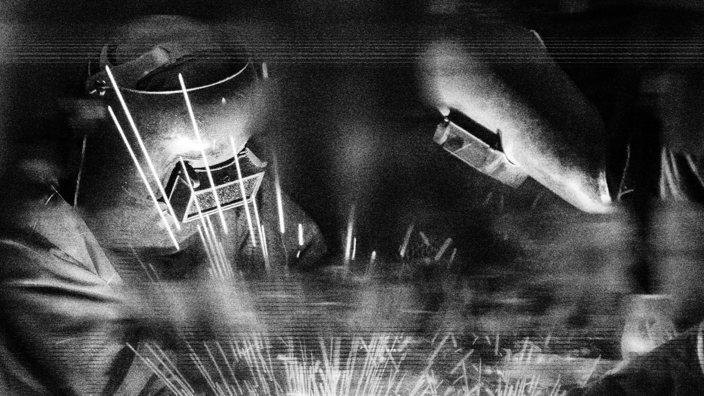 Zwei Menschen mit Schweißermaske sind bei der Arbeit. Schwarz-weiß Optik.