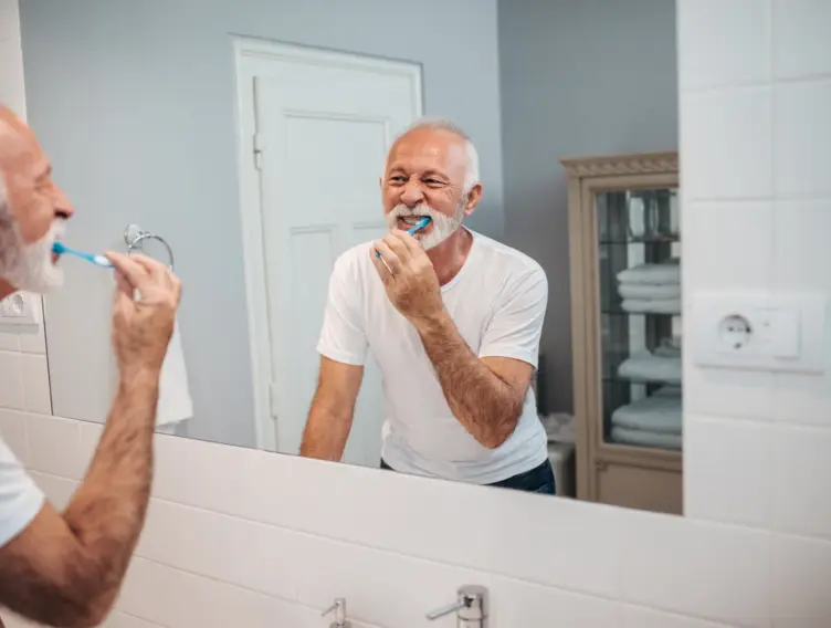 Älterer Mann steht vor dem Badezimmerspiegel und putzt sich die Zähne.
