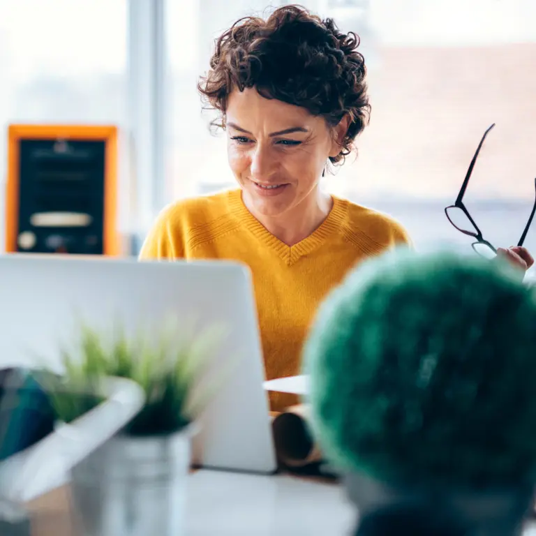 Frau sitzt lächelnd im Büro am Schreibtisch vor einem Laptop. 