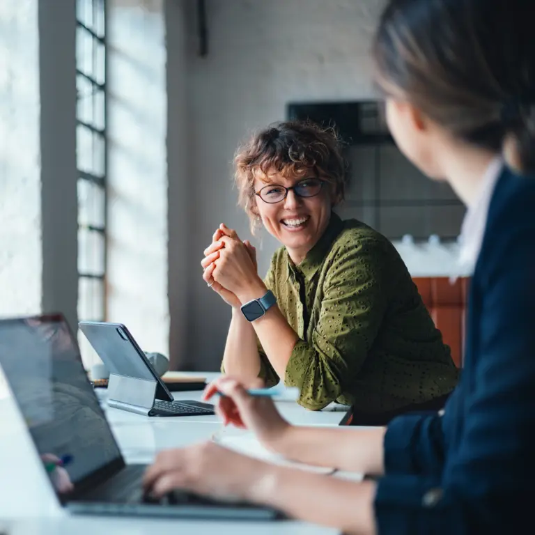 Zwei Frauen sitzen im Büro und am Schreibtisch und lächeln sich an. 