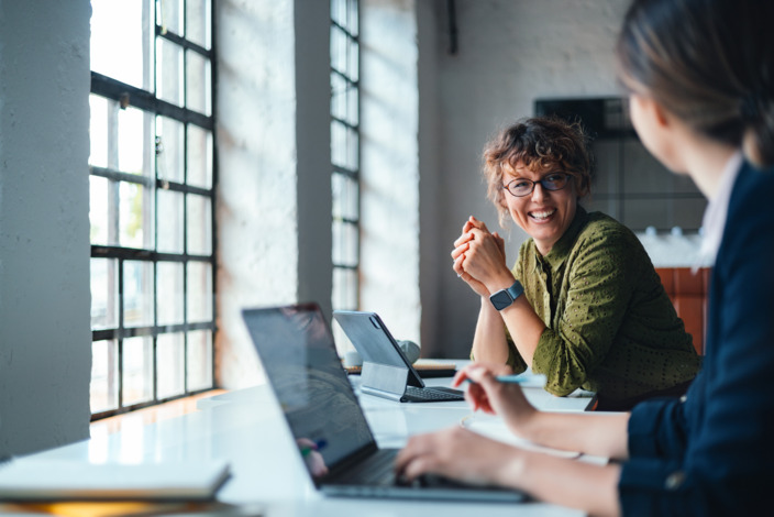 Zwei Frauen sitzen im Büro und am Schreibtisch und lächeln sich an. 