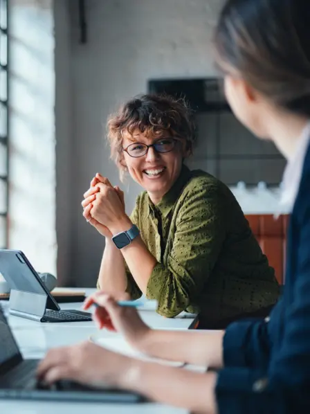 Zwei Frauen sitzen im Büro und am Schreibtisch und lächeln sich an. 