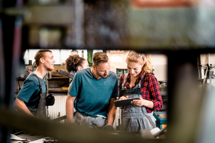 Eine Gruppe Handwerker steht in einer Werkstatt und schaut auf ein Tablet. 