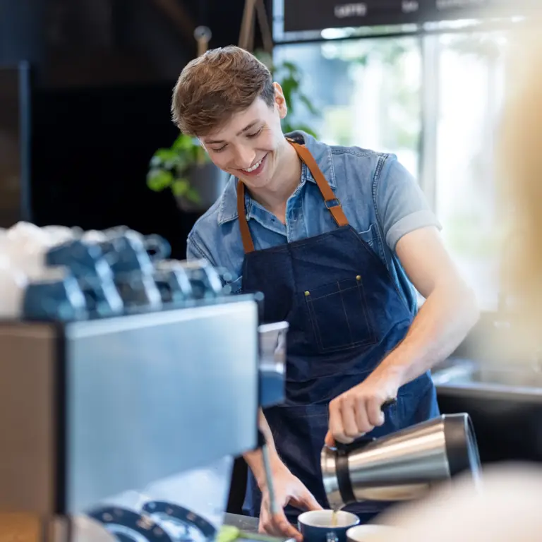 Student steht in einem Cafe und bereitet einen Kaffeegetränk zu. 