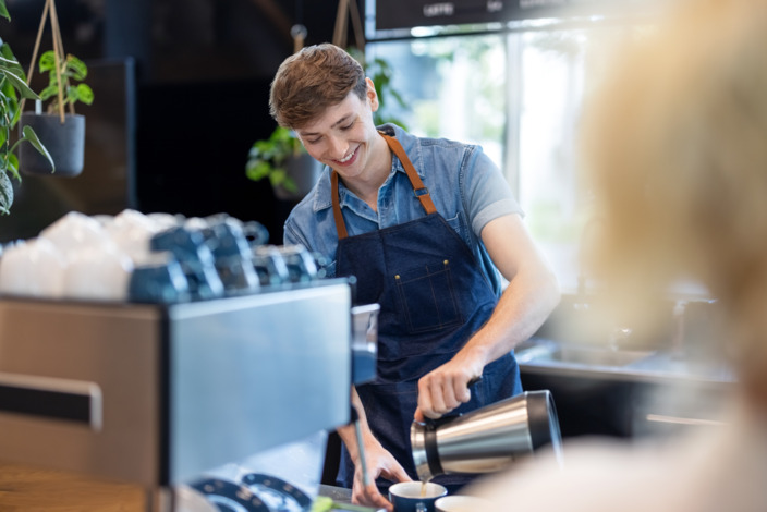 Student steht in einem Cafe und bereitet einen Kaffeegetränk zu. 