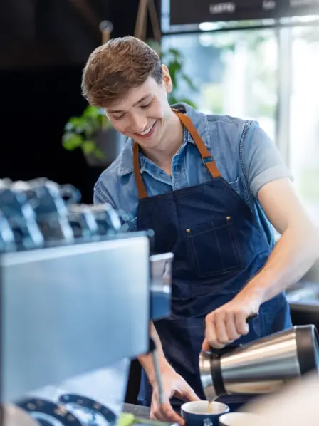 Student steht in einem Cafe und bereitet einen Kaffeegetränk zu. 