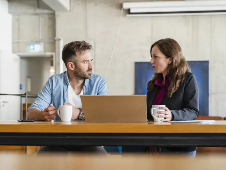 Mann und Frau sitzen mit einem Laptop am Tisch und unterhalten sich.