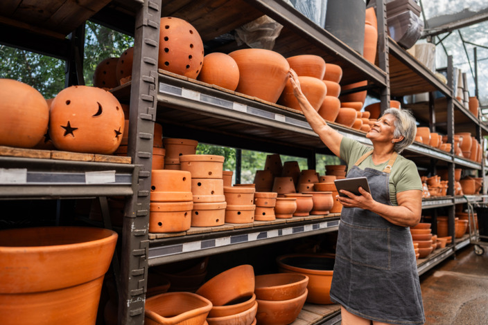 Ältere Frau arbeitet in einem Baumarkt und steht vor einem Blumentopf-Regal.
