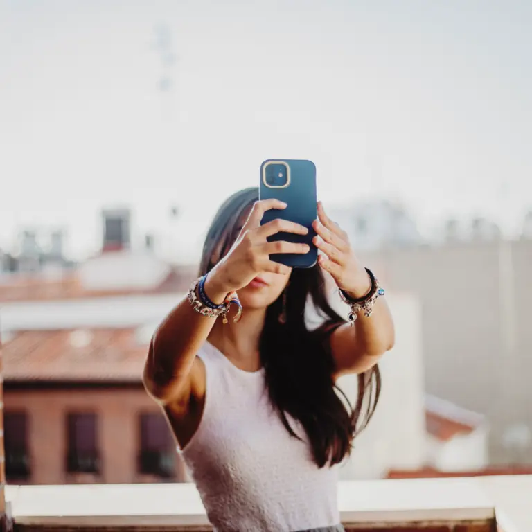 Junge Frau mit braunen Haaren steht auf einem Dach und macht ein Selfie mit ihrem Smartphone.