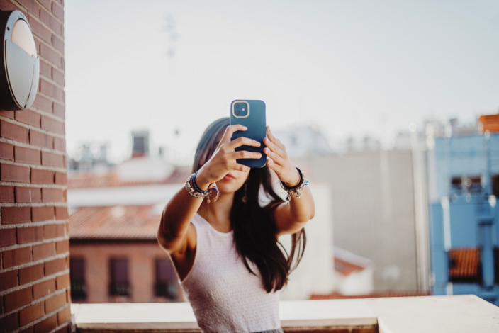 Junge Frau mit braunen Haaren steht auf einem Dach und macht ein Selfie mit ihrem Smartphone.