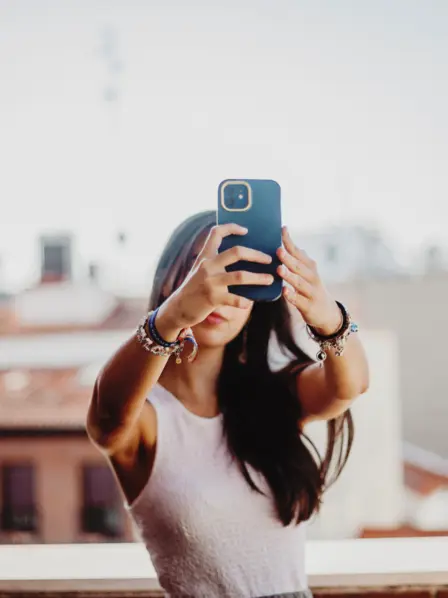 Junge Frau mit braunen Haaren steht auf einem Dach und macht ein Selfie mit ihrem Smartphone.