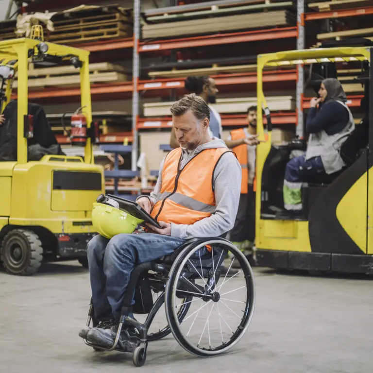 Mann im Rollstuhl trägt eine Warnweste und arbeitet mit anderen Personen zusammen im Lager.