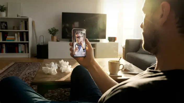 Mann sitzt in seinem Wohnzimmer und hat einen Videocall mit einem Arzt auf seinem Handy.