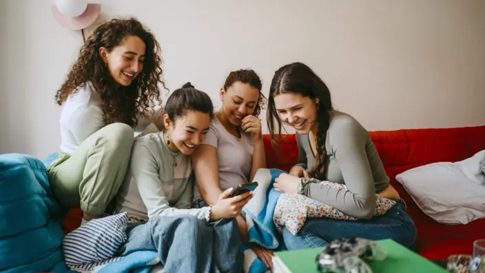 Vier junge Frauen sitzen auf dem Sofa und schauen lachend auf ein Smartphone.