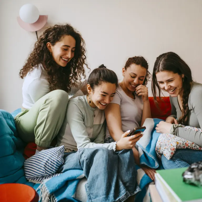 Vier junge Frauen sitzen auf dem Sofa und schauen lachend auf ein Smartphone.