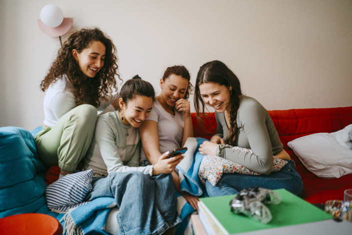 Vier junge Frauen sitzen auf dem Sofa und schauen lachend auf ein Smartphone.