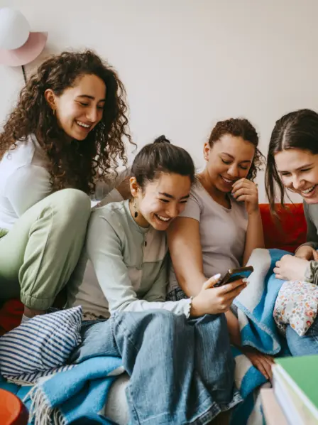 Vier junge Frauen sitzen auf dem Sofa und schauen lachend auf ein Smartphone.
