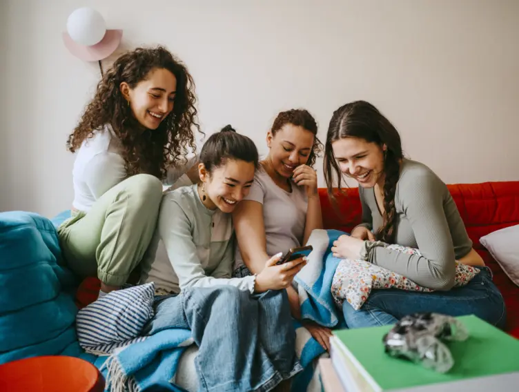 Vier junge Frauen sitzen auf dem Sofa und schauen lachend auf ein Smartphone.
