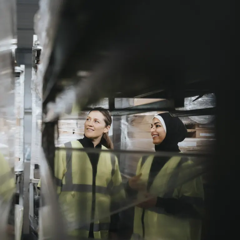 Zwei Arbeiterinnen stehen mit Warnwesten in einer Lagerhalle.