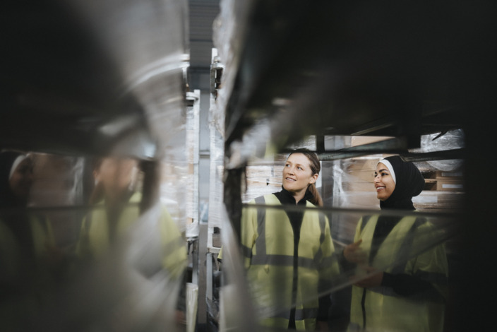 Zwei Arbeiterinnen stehen mit Warnwesten in einer Lagerhalle.