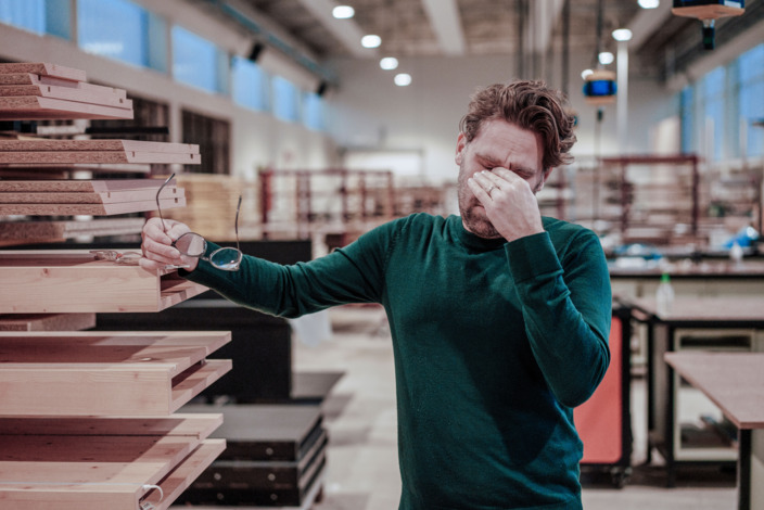 Mann steht in der Handwerkshalle und hält sich die Hand an die Augen-Nasen-Patie während er sich mit der anderen Hand abstützen muss.