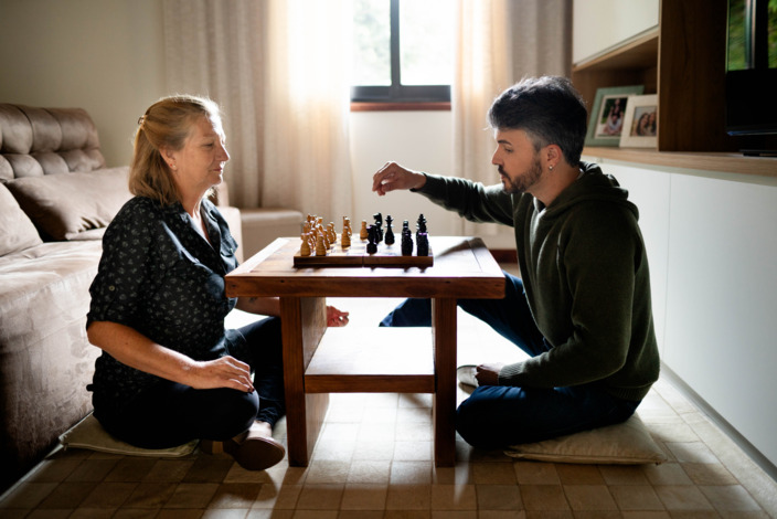 Ältere Frau spielt mit jüngerem Mann am Wohnzimmertisch Schach.
