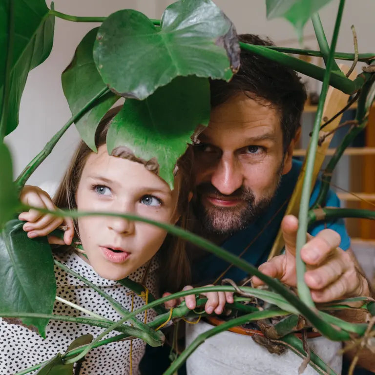 Vater und Tochter spielen hinter einen Zimmerpflanze, als seien sie im Dschungel.