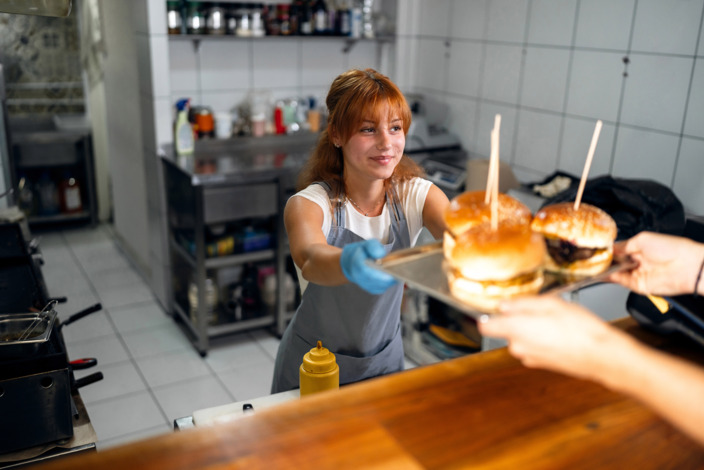 Frau steht im Imbiss und übergibt ein Tablett mit Burgern an einen Kunden. 