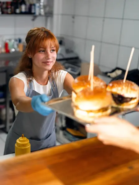 Frau steht im Imbiss und übergibt ein Tablett mit Burgern an einen Kunden. 