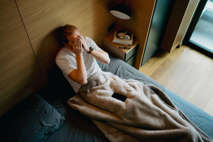 Mann sitzt im Bett und reibt seine Hände im Gesicht.