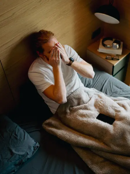 Ein Mann sitzt im Bett und reibt sich mit seinen Händen übers Gesicht.