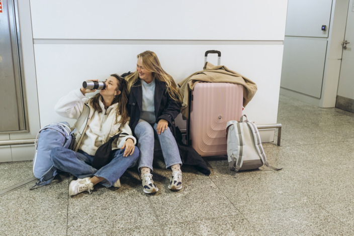 Zwei junge Frauen sitzen lachend mit Gepäck auf dem Boden am Flughafen.