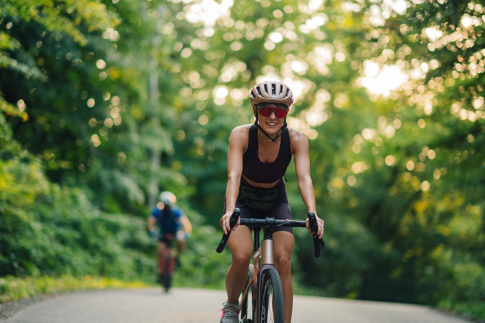 Fahrradfahrerin fährt in Sportkleidung auf einer geteerten Straße durch den Wald.