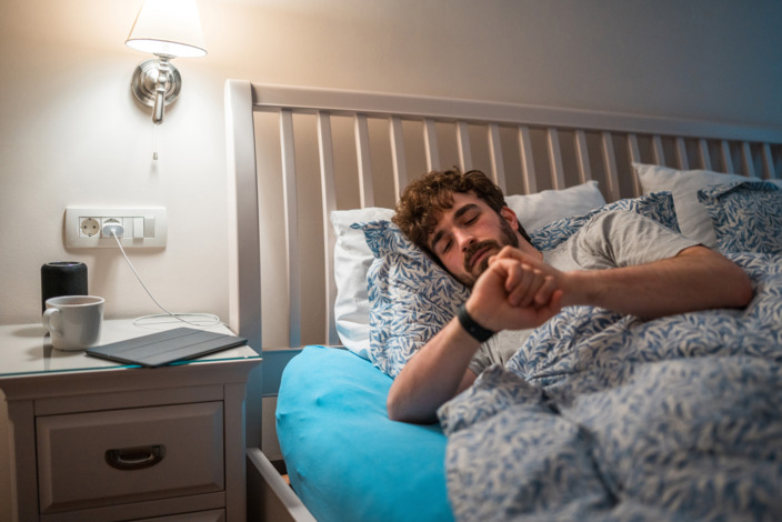 Mann liegt in einem Schlafzimmer im Bett und schaut auf seine Smartwatch. 