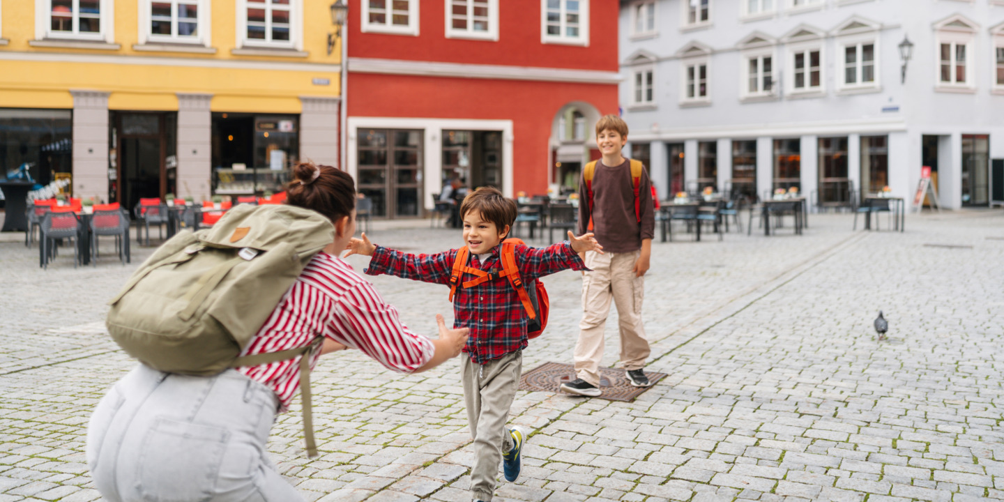 Mutter steht gebeugt mit offenen Armen auf einem Markplatz und empfängt ihre Schulkinder.