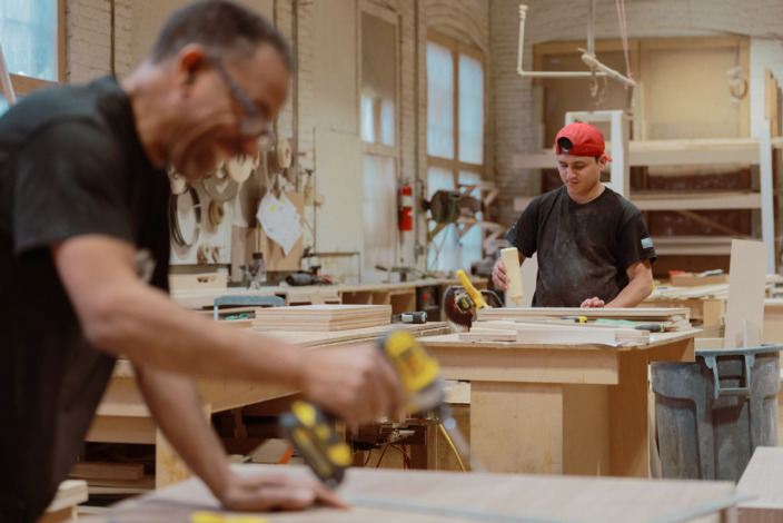 Schreiner und Auszubildender arbeiten mit Holz in der Werkstatt.