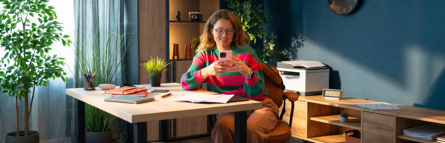 Frau sitzt am Schreibtisch und schaut aufs Handy.