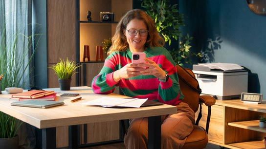 Frau sitzt mit ihrem Handy an einem Schreibtisch.