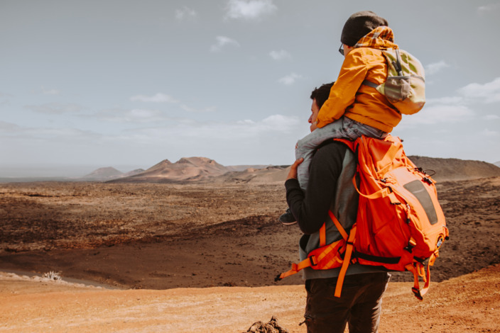 Vater im Trekking-Outfit trägt seinen Sohn auf den Schultern und blickt in eine Wüstenlandschaft