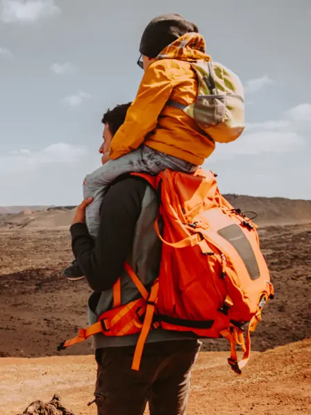 Vater im Trekking-Outfit trägt seinen Sohn auf den Schultern und blickt in eine Wüstenlandschaft