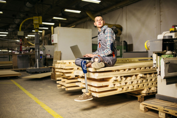 junger Handwerker mit einer Beinprothese sitzt auf einem Bretterstapel in der Werkstatt