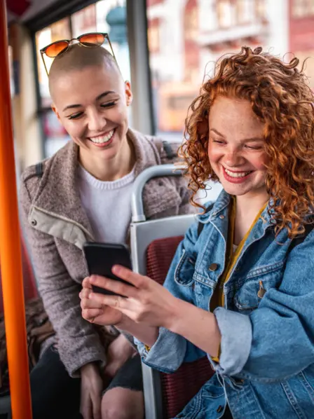Zwei junge Frauen sitzen im Bus und schauen lachend auf ein Smartphone.