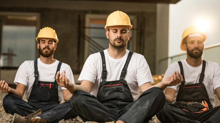 Drei Bauarbeiter sitzen nebeneinander im Schneidersitz und meditieren.