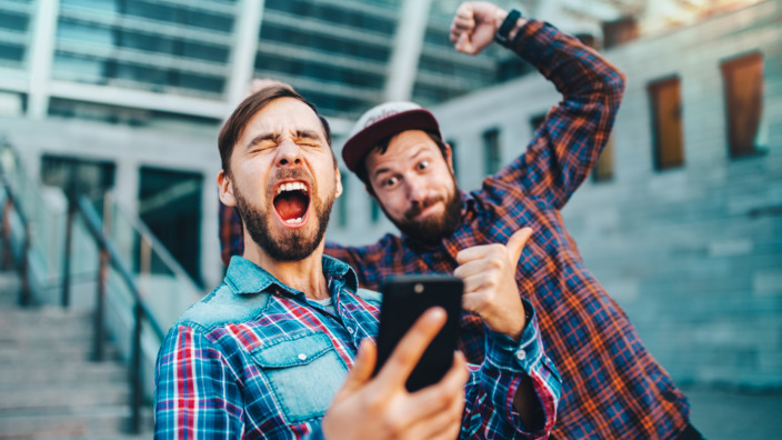 zwei junge Männer schauen auf ein Smartphone und jubeln