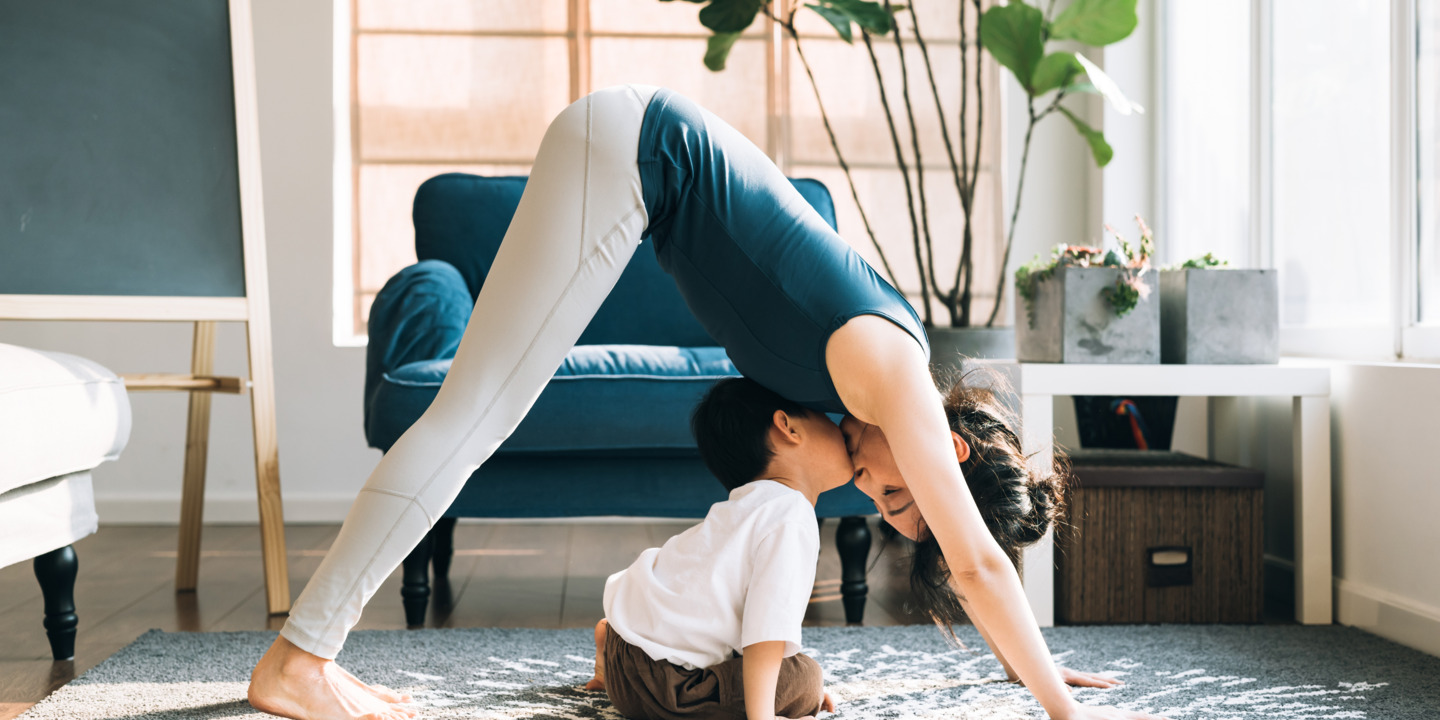 Frau absolviert die Yoga-Übung herabschauender Hund, ihr kleiner Sohn sitzt zwischen ihren Armen und Beinen