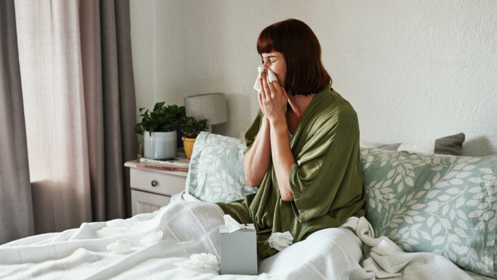 Junge Frau sitzt auf dem Bett und niest in Taschentuch