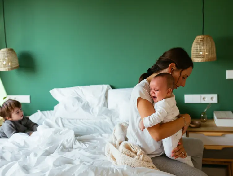 Mutter sitzt erschöpft auf dem Bett und tröstet ihr weinendes Baby, der zweijährige Bruder schaut zu